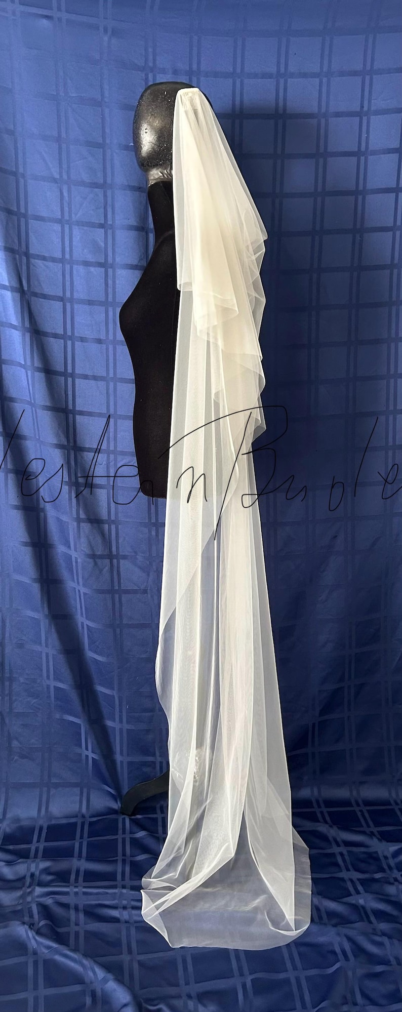 Puede incluir: Un velo de novia blanco con m&uacute;ltiples capas de tela suave y transparente. El velo est&aacute; drapeado sobre una cabeza y hombros de maniqu&iacute; negro, mostrando su dise&ntilde;o fluido. El fondo es una tela azul.