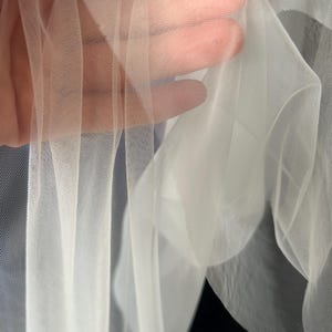 Puede incluir: Primer plano de tela blanca transparente, posiblemente tul o red. La tela se sostiene, revelando su delicada textura transparente. El material parece suave y fluido, con un ligero brillo.