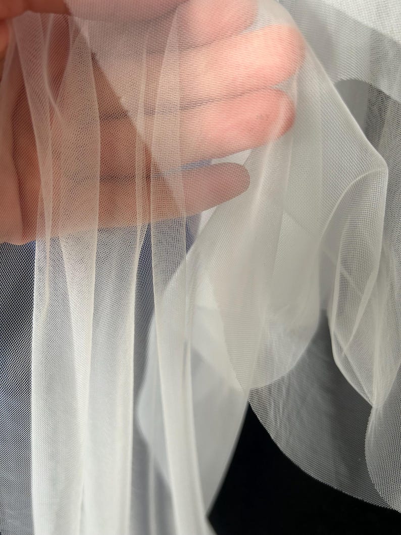 Puede incluir: Primer plano de tela blanca transparente, posiblemente tul o red. La tela se sostiene, revelando su delicada textura transparente. El material parece suave y fluido, con un ligero brillo.