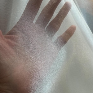 Op de afbeelding: Close-up van een doorschijnende, gebroken witte stof. De stof wordt omhoog gehouden, waardoor licht erdoorheen kan schijnen, waardoor de textuur en doorschijnendheid zichtbaar worden. Het materiaal lijkt licht en delicaat, mogelijk voor kleding of decoratief gebruik.