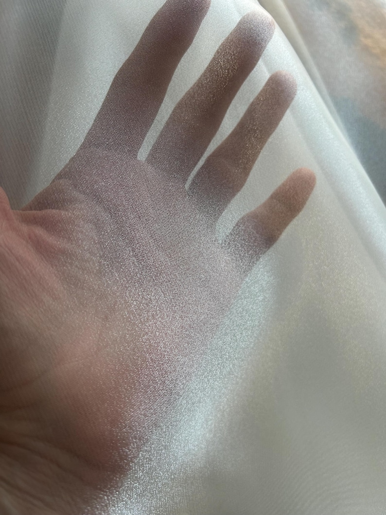 Op de afbeelding: Close-up van een doorschijnende, gebroken witte stof. De stof wordt omhoog gehouden, waardoor licht erdoorheen kan schijnen, waardoor de textuur en doorschijnendheid zichtbaar worden. Het materiaal lijkt licht en delicaat, mogelijk voor kleding of decoratief gebruik.