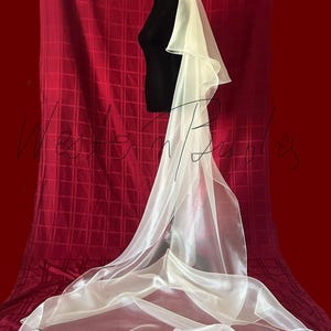 Op de afbeelding: Een lange, vloeiende ivoorkleurige bruidssluier. De sluier is gemaakt van een transparante, lichtgewicht stof en is over een zwarte mannequin gedrapeerd. De sluier valt naar beneden en cre&euml;ert zachte plooien en een delicate uitstraling. De achtergrond is een dieprode stof.
