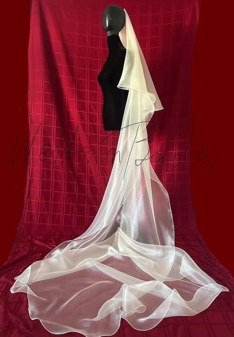 Op de afbeelding: Een lange, vloeiende ivoorkleurige bruidssluier. De sluier is gemaakt van een transparante, lichtgewicht stof en is over een zwarte mannequin gedrapeerd. De sluier valt naar beneden en cre&euml;ert zachte plooien en een delicate uitstraling. De achtergrond is een dieprode stof.