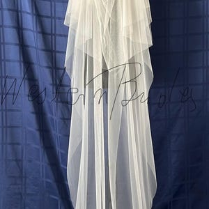 Puede incluir: Un velo de novia blanco y transparente con m&uacute;ltiples capas, colocado sobre un maniqu&iacute;. El velo es largo y fluido, con una textura suave y delicada. El fondo es una tela azul oscuro con un patr&oacute;n de cuadr&iacute;cula.