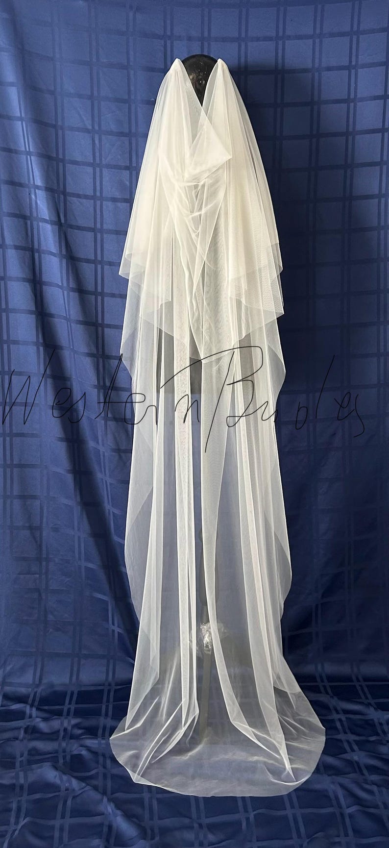 Puede incluir: Un velo de novia blanco y transparente con m&uacute;ltiples capas, colocado sobre un maniqu&iacute;. El velo es largo y fluido, con una textura suave y delicada. El fondo es una tela azul oscuro con un patr&oacute;n de cuadr&iacute;cula.