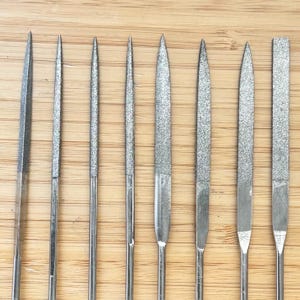 Könnte beinhalten: Ein Satz von neun Metall-Nadelfeilen in verschiedenen Formen und Größen. Die Feilen haben eine silberne Oberfläche und sind vertikal auf einer hellbraunen Holzoberfläche angeordnet. Die Spitzen der Feilen sind spitz oder flach.
