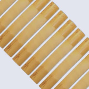 May include: A collection of light brown reeds, likely for a musical instrument, arranged diagonally against a white background. The reeds have a cylindrical shape with darker brown bands and tips, suggesting a natural material.
