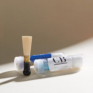 May include: A wooden oboe reed with a black and gold ferrule, next to two small white tubes with blue labels. The labels read "WOODWINDS" and "CB Fine Quality Woodwinds".