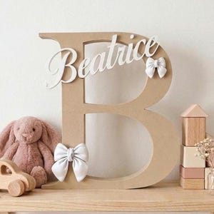 May include: A large, tan, free-standing letter B made of a smooth, matte material. The name Beatrice is written in white cursive script across the top section. Two white fabric bows are attached to the letter, one at the bottom left and one on the right side.
