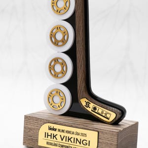 Puede incluir: Trofeo de hockey con base de madera y placa dorada. El trofeo presenta un palo de hockey y cuatro discos blancos y dorados. La placa dice "IHK VIKINGI".