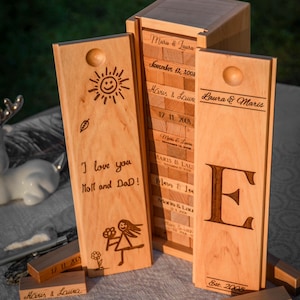 May include: A wooden game set with a personalized message, "I love you Moll and Dad!" and a drawing of a girl with a flower. The game set is made of natural wood and has a light brown color. The game set is in a box with a lid that has the names "Maris & Laura" and the date "November 12, 2008" engraved on it. The box also has a side panel with the letter "E" engraved on it.