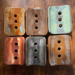 May include: Six rectangular ceramic soap dishes arranged in two rows. Each dish features a glossy, multi-coloured glaze and three circular drainage holes in the centre. The colours include shades of brown, orange, teal, and dark grey with variations.