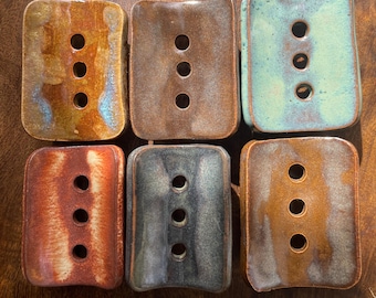 Puede incluir: Seis jaboneras rectangulares de cerámica dispuestas en dos filas. Cada pieza presenta un esmalte brillante multicolor y tres orificios de drenaje circulares en el centro. Los colores incluyen tonos de marrón, naranja, verde azulado y gris oscuro.