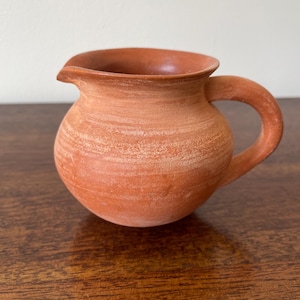 May include: A small, round terracotta jug with a matte, earthy orange finish. The vessel features a wide, flared rim, a distinct pouring spout, and a curved handle attached to the side. The surface shows subtle horizontal texture lines.