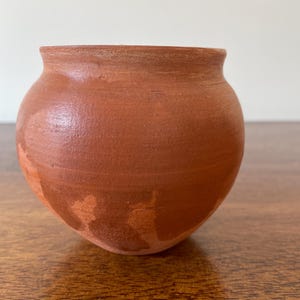May include: A small, round terracotta pot with a wide, open mouth and a smooth, matte finish. The vessel features a natural, earthy reddish-brown colour with subtle variations in tone across its surface, showcasing a simple, unadorned design.