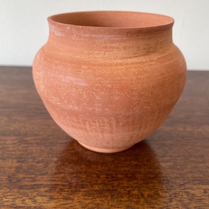 May include: A small, round terracotta pot with a wide opening and a slightly flared rim. The exterior surface features a natural, matte, reddish-brown finish with visible horizontal texture lines from the crafting process and a rounded base.