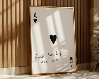 May include: A gold framed print with a white background and black text that reads "how lucky are we". The print features a black ace of spades playing card with a black heart in the center.