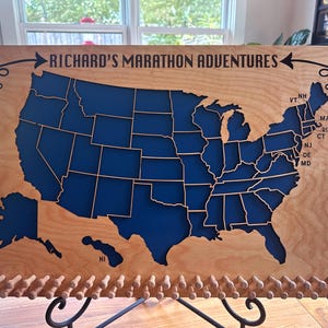 May include: A wooden map of the United States with a blue painted outline of each state. The map is titled "Richard's Marathon Adventures" and has a row of wooden pegs at the bottom.
