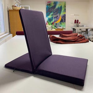 Può includere: Un cuscino da meditazione pieghevole di colore viola con una cerniera. Il cuscino è piegato in posizione seduta.
