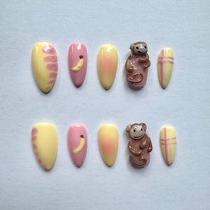 May include: A set of ten almond-shaped false nails. The nails feature a variety of designs, including yellow and pink gradients, banana decals, monkey figurines, and yellow nails with pink plaid patterns. The nails are arranged on a light blue surface.