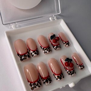 May include: A set of ten false nails with a leopard print design and a red accent. Each nail has a cherry on top.