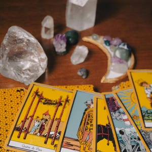 May include: A collection of tarot cards with yellow borders and illustrations, alongside various crystals and a crescent moon-shaped decorative piece. The cards feature images and text in a foreign language. The items are arranged on a wooden surface.