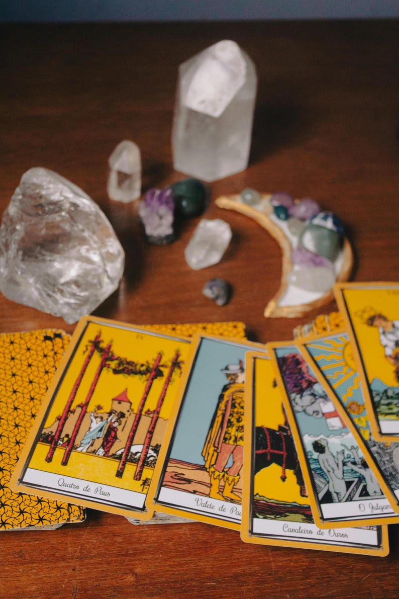 May include: A collection of tarot cards with yellow borders and illustrations, alongside various crystals and a crescent moon-shaped decorative piece. The cards feature images and text in a foreign language. The items are arranged on a wooden surface.