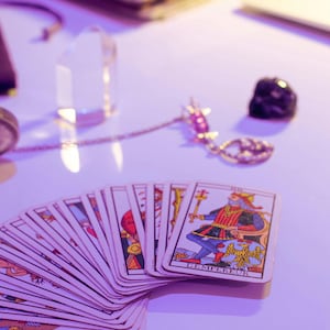 May include: A deck of tarot cards fanned out on a white surface, with the card "L'Empereur" visible. The cards feature colorful illustrations. A clear crystal, a gold-colored necklace with a pendant, and a black stone are also present.