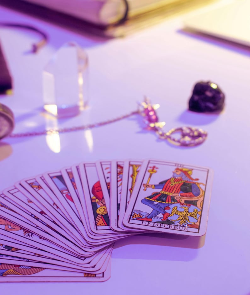 May include: A deck of tarot cards fanned out on a white surface, with the card "L'Empereur" visible. The cards feature colorful illustrations. A clear crystal, a gold-colored necklace with a pendant, and a black stone are also present.