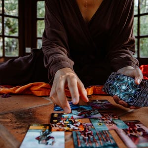 May include: A person is seen arranging tarot cards on a terracotta tile surface. The cards feature colorful illustrations. The person is wearing a brown wrap top and holding a deck of cards with a blue and silver design.