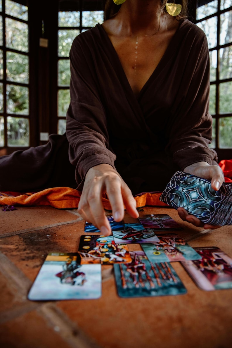 May include: A person is seen arranging tarot cards on a terracotta tile surface. The cards feature colorful illustrations. The person is wearing a brown wrap top and holding a deck of cards with a blue and silver design.