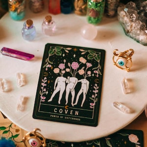 May include: A collection of mystical items including a tarot card with the word "COVEN" and the phrase "POWER OF SISTERHOOD", small glass bottles, crystals, and a gold ring with an eye design. The items are arranged on a white surface.
