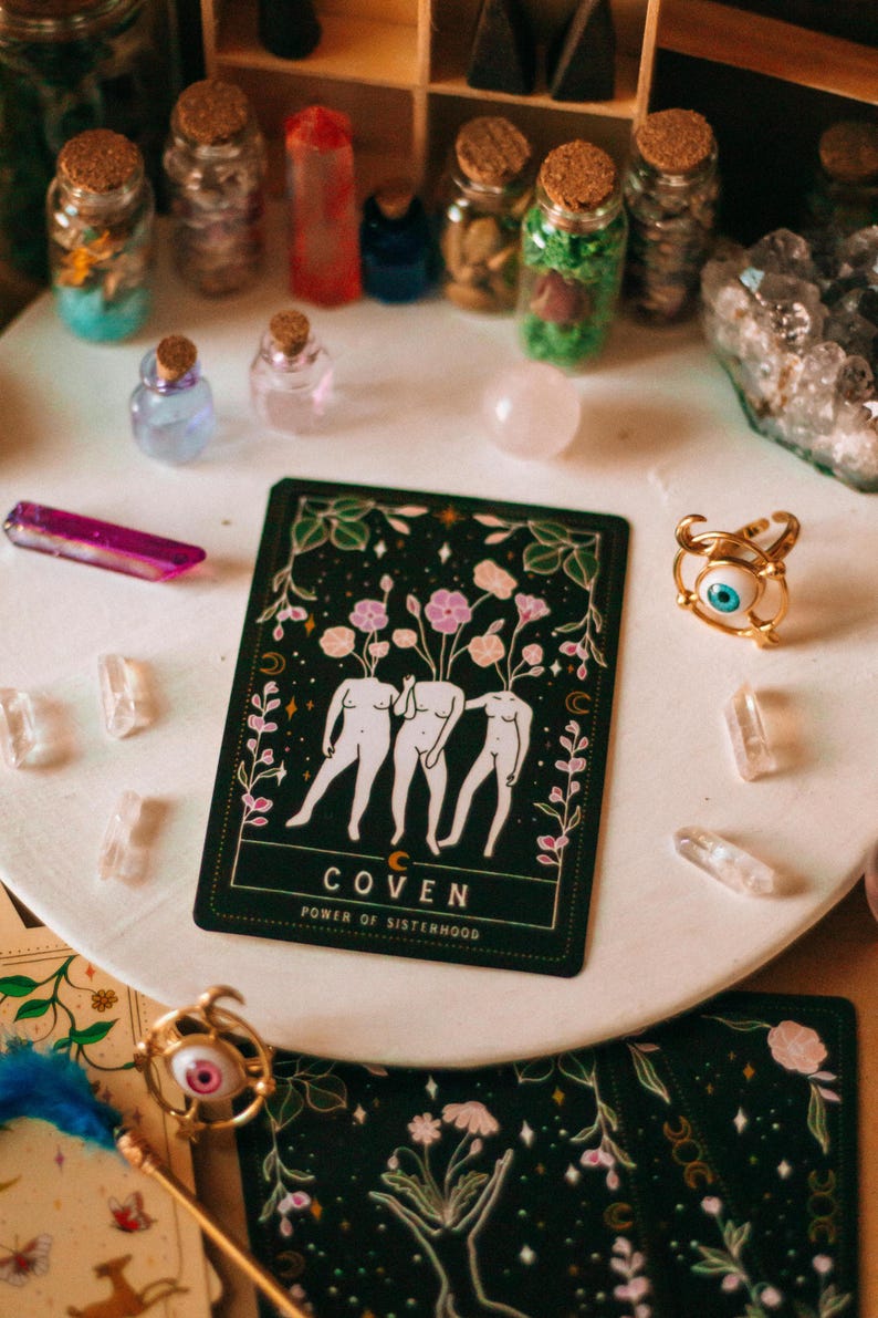 May include: A collection of mystical items including a tarot card with the word "COVEN" and the phrase "POWER OF SISTERHOOD", small glass bottles, crystals, and a gold ring with an eye design. The items are arranged on a white surface.