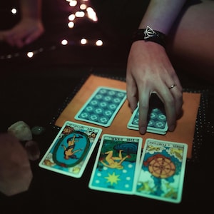 May include: A tarot card reading setup with several cards laid out on a tan surface. The cards feature colorful illustrations and titles. A hand is reaching for a deck of cards. Crystals and fairy lights are also visible.