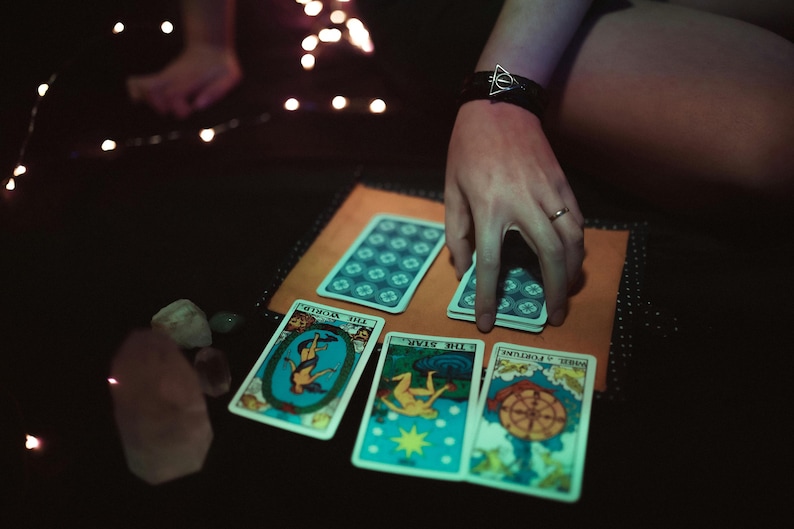 May include: A tarot card reading setup with several cards laid out on a tan surface. The cards feature colorful illustrations and titles. A hand is reaching for a deck of cards. Crystals and fairy lights are also visible.
