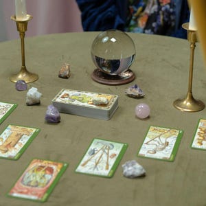 May include: A divination scene featuring a crystal ball on a wooden stand, tarot cards, and various crystals. Two brass candle holders with lit candles flank the scene. The cards are spread out on a green surface.