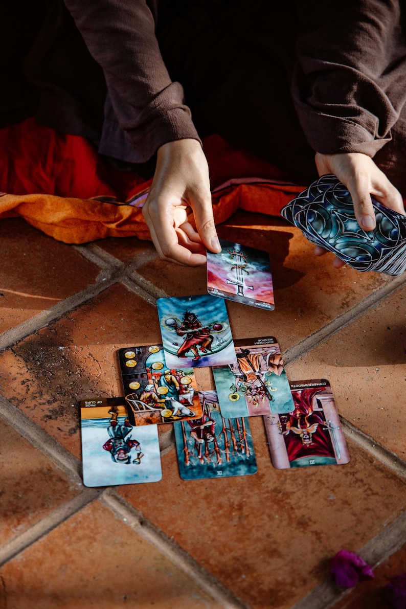 May include: A collection of tarot cards with colorful illustrations, including images of figures, symbols, and scenes. The cards are arranged on a terracotta tile surface, with hands holding and selecting cards from the deck. The cards have a mystical theme.