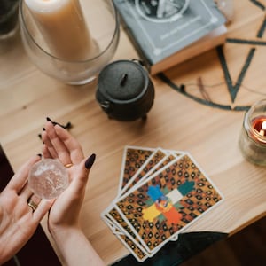 May include: A crystal ball held in hands, with tarot cards, a candle, and a small cauldron on a wooden table. A lit candle sits in a glass jar. A book and a large candle are also visible.