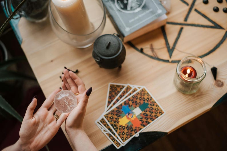 May include: A crystal ball held in hands, with tarot cards, a candle, and a small cauldron on a wooden table. A lit candle sits in a glass jar. A book and a large candle are also visible.