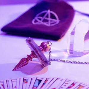 May include: A divination pendulum with a brass-colored metal body and a clear, circular crystal. A chain is attached to the pendulum. A deck of tarot cards is fanned out in the foreground. A dark purple pouch with a white symbol is in the background.