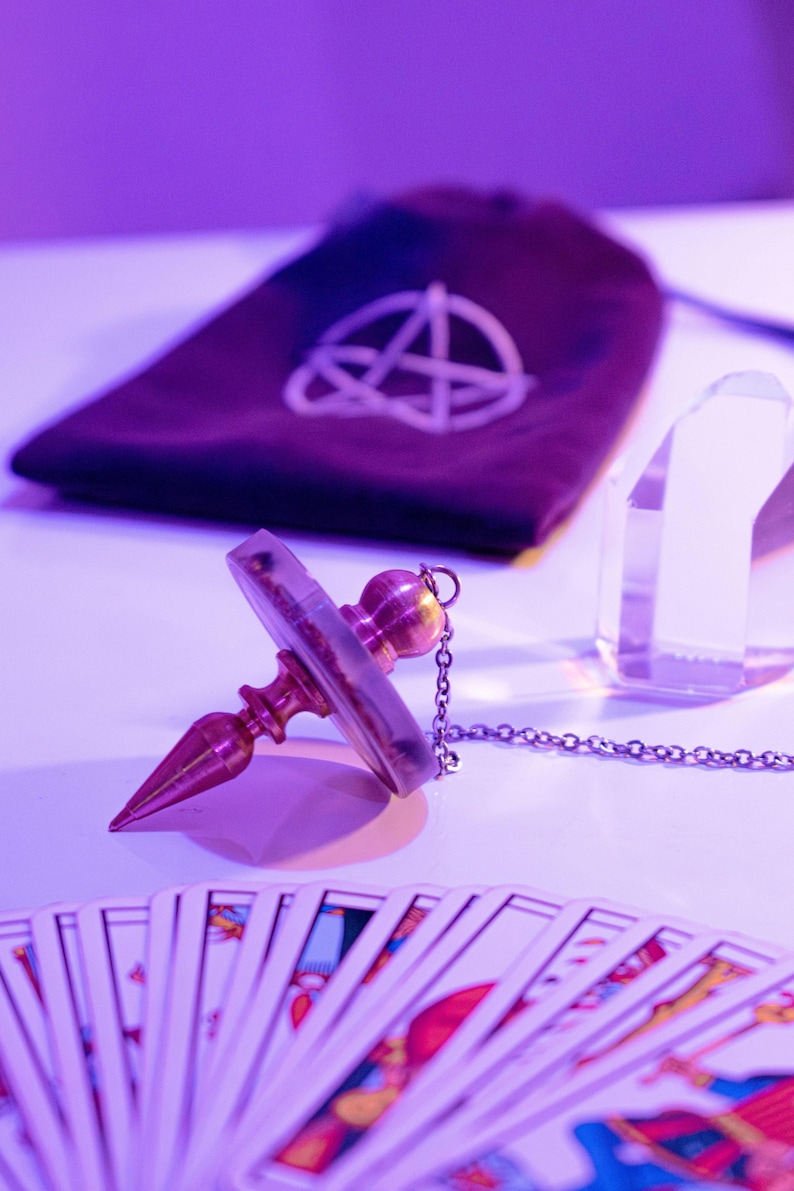 May include: A divination pendulum with a brass-colored metal body and a clear, circular crystal. A chain is attached to the pendulum. A deck of tarot cards is fanned out in the foreground. A dark purple pouch with a white symbol is in the background.