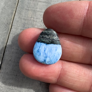 Könnte beinhalten: Ein tropfenförmiger Stein mit einem blau-schwarzen Muster. Der obere Teil ist dunkel mit schwarzen und grünen Streifen, während der untere Teil hellblau mit Marmoreffekt ist. Der Stein wird zwischen Fingern gehalten.
