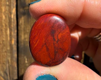 May include: An oval-shaped polished gemstone with a smooth, glossy surface. The stone features a vibrant red base color with intricate, dark brown to black vein-like patterns running throughout the surface, creating a marbled appearance.