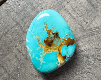 May include: A polished, oval-shaped turquoise gemstone with a vibrant blue hue. The stone features intricate brown and gold matrix patterns, creating a unique and natural design. The gemstone is set against a gray surface.