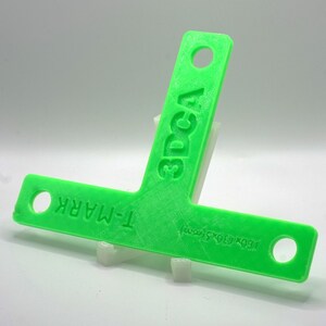 May include: A vibrant green T-square featuring the text "T-MARK" and "3DCA". It has holes at each end and is supported by a white stand. The dimensions are 160 x 130.5 mm.