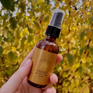 May include: A brown glass bottle with a gold label that reads "Radiant Essential Oil Spray" and "Sacred Space Rituals". The bottle has a black spray top and is held in a hand.