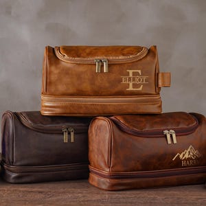 Könnte beinhalten: Drei braune Kulturbeutel aus Leder mit Reißverschlüssen sind gestapelt. Die obere Tasche ist hellbraun und trägt den Namen "ELLIOT". Die mittlere Tasche ist dunkelbraun. Die untere Tasche ist braun und hat eine Berg-Grafik und den Namen "HARRY".