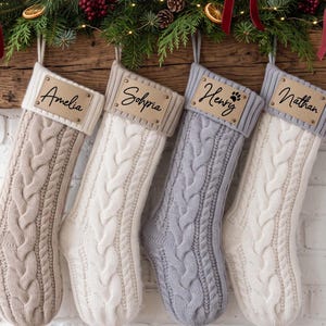 Könnte beinhalten: Vier Weihnachtsstrümpfe im Zopfstrickmuster in Beige, Creme und Grau. Jeder Strumpf hat ein geripptes Bündchen mit einem rechteckigen Lederetikett, das mit zwei Metallnieten befestigt ist. Die Etiketten sind mit den Namen Amelia, Sophia, Henry und Nathan versehen.
