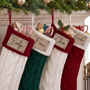 Könnte beinhalten: Vier Weihnachtsstrümpfe aus Zopfstrick in Creme, Grün und Rot. Jeder Strumpf hat ein personalisiertes, rechteckiges Namensschild aus Lederimitat mit den Namen Evelyn, Fiona, Claire und Amelia, gefüllt mit kleinen Geschenkboxen und Zuckerstangen.
