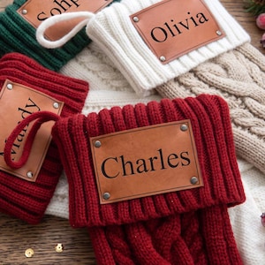 Könnte beinhalten: Ein Set aus vier gestrickten Weihnachtsstrümpfen in Rot, Weiß, Grün und Beige, jeweils mit einem rechteckigen braunen Lederaufnäher, der mit Metallnieten befestigt ist. Die Aufnäher sind mit den Namen Sohpia, Olivia, Mandy und Charles graviert.